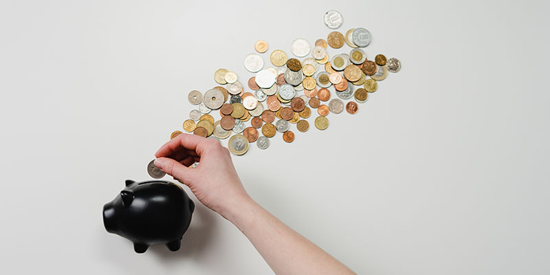 A hand placing a coin into a black piggy bank with several coins scattered on a white surface