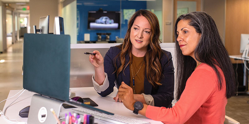 Two business women talking about sales in office at desk with laptop