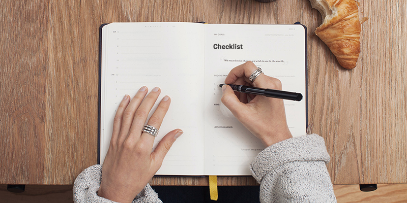 A woman writing a checklist in a black notebook on a wooden desk
