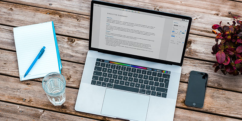 A laptop, notebook, glass of water, pen, and cellphone on a brown wood table