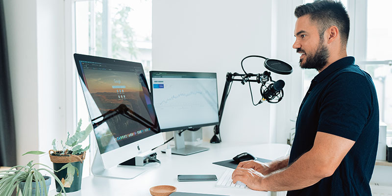 Man at work with podcast microphone in front of a desk and two screens with splitscreen computer and laptop on the desk in a social media agency.